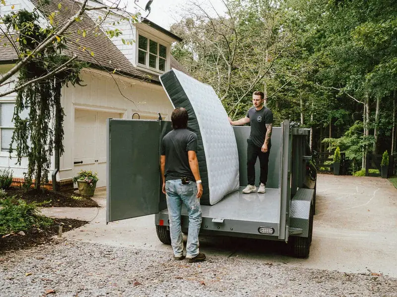 Two-person crew loading a mattress into a dump trailer in a driveway